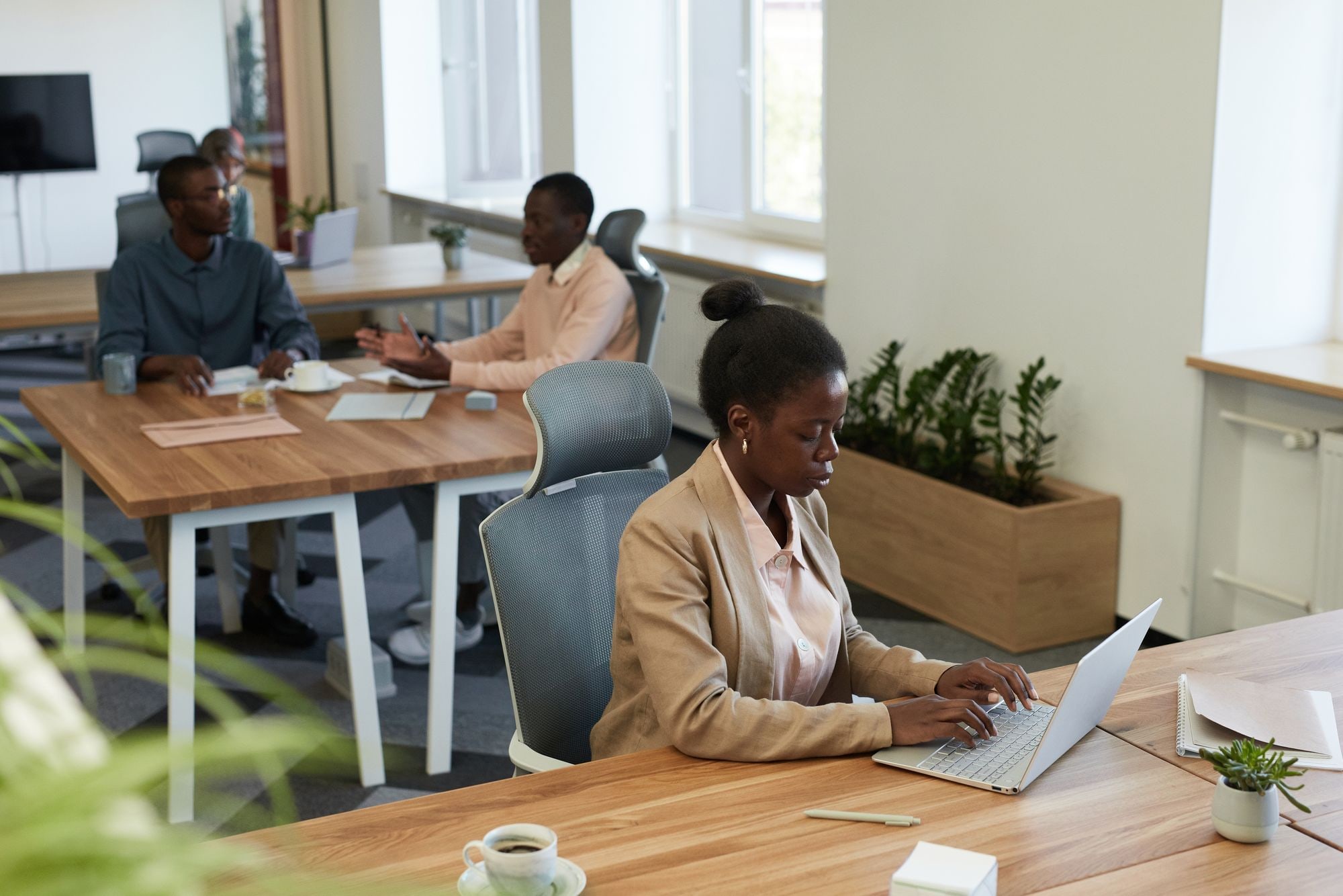 Woman using a laptop in a modern office while colleagues discuss work at a table nearby
